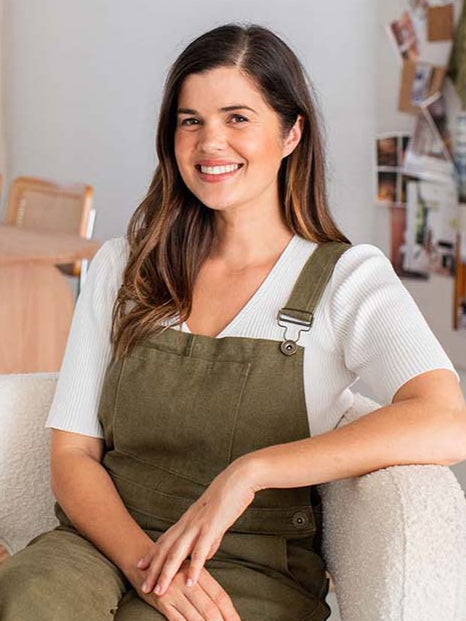 Woman sitting on a couch wearing a white shirt and green overalls, with a home interior background.