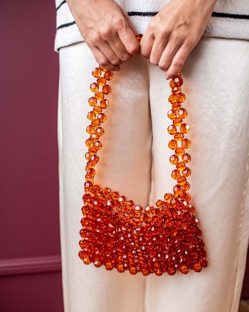 A person wearing cream-colored trousers holds a small shoulder bag made of faceted amber-orange glass beads. The background features a dark burgundy wall and a wooden floor.