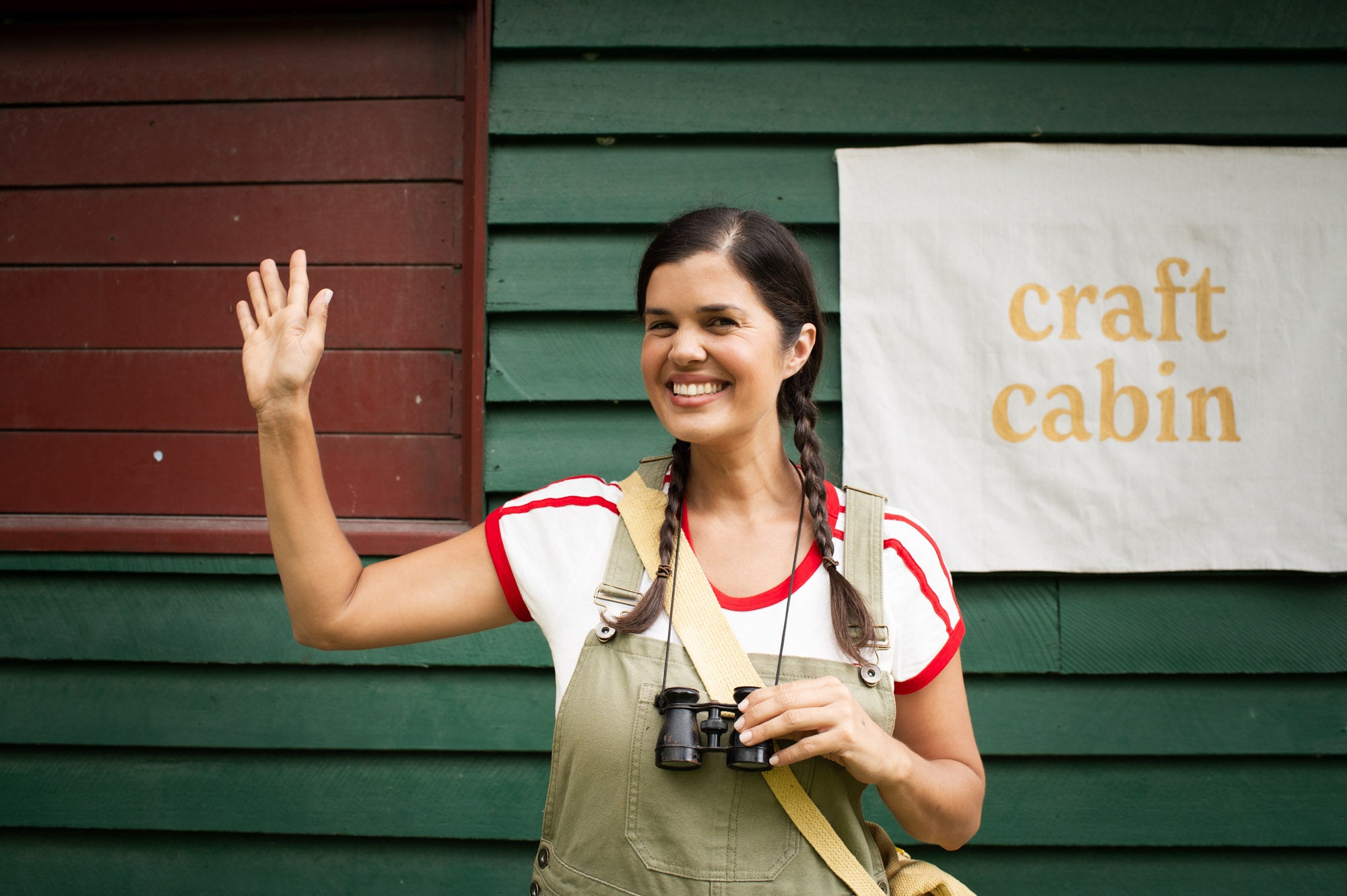 Woman waving in front of a green cabin with a 'craft cabin' sign.